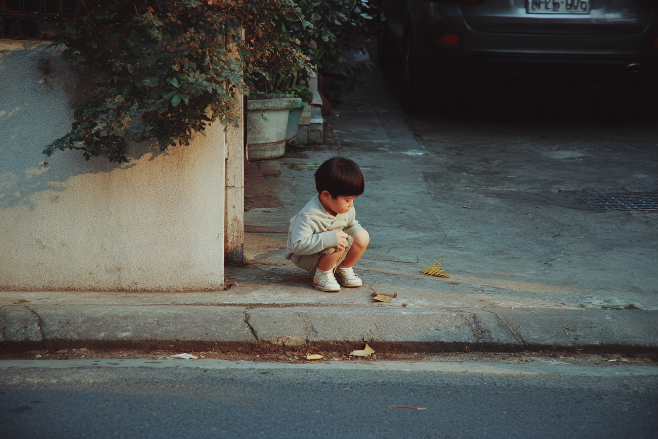 Toddler crouching to examine a crack and leaf on the pavement during the walk home while a parent waits nearby.