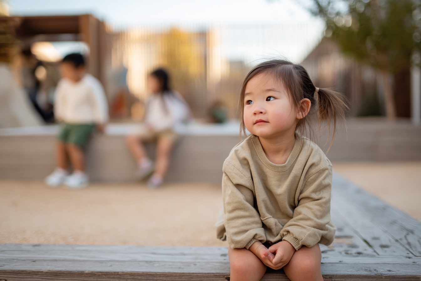 Toddler sitting quietly and observing other children playing from a short distance.