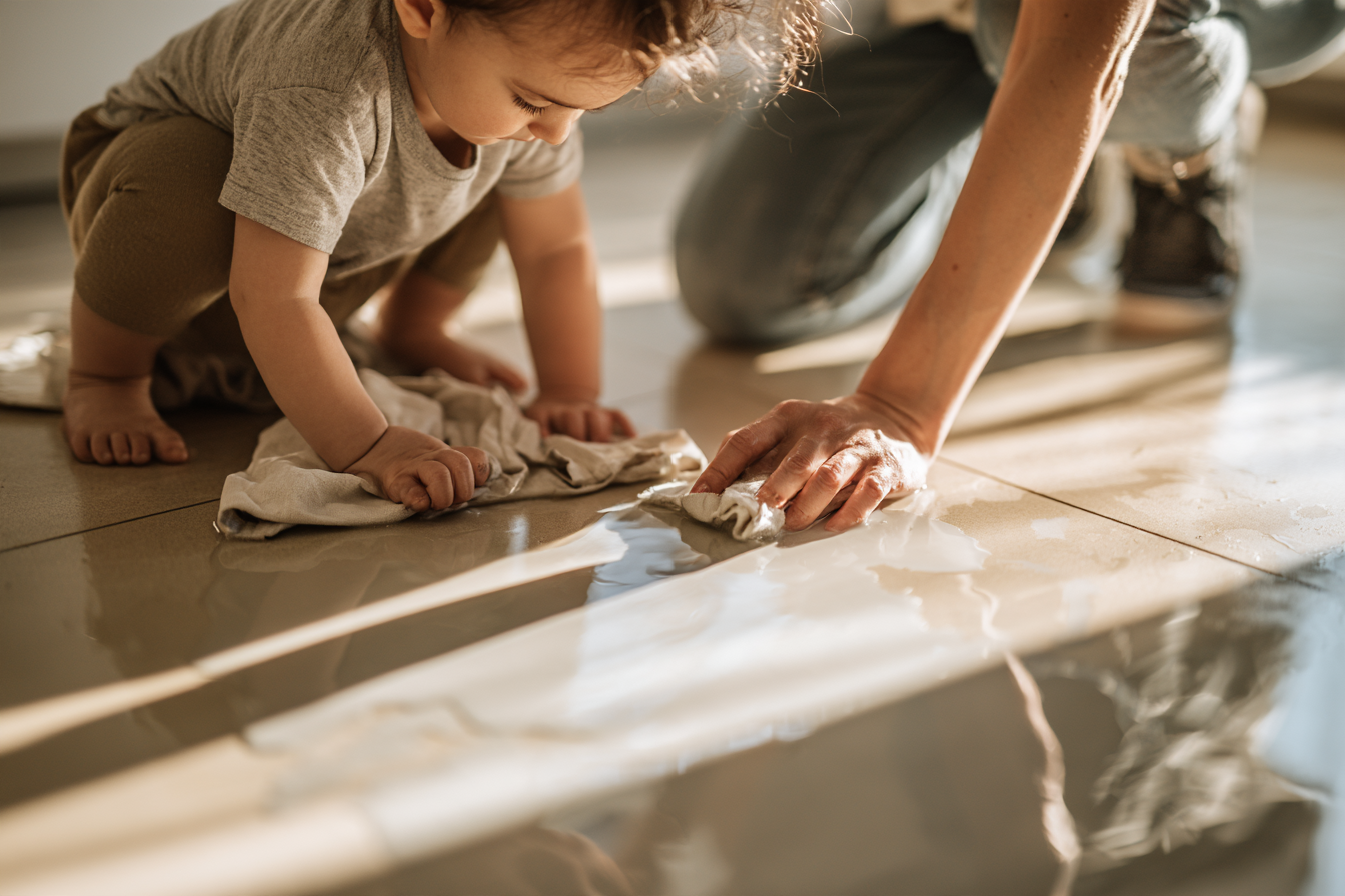 A toddler and adult using cloths to wipe water from a kitchen floor